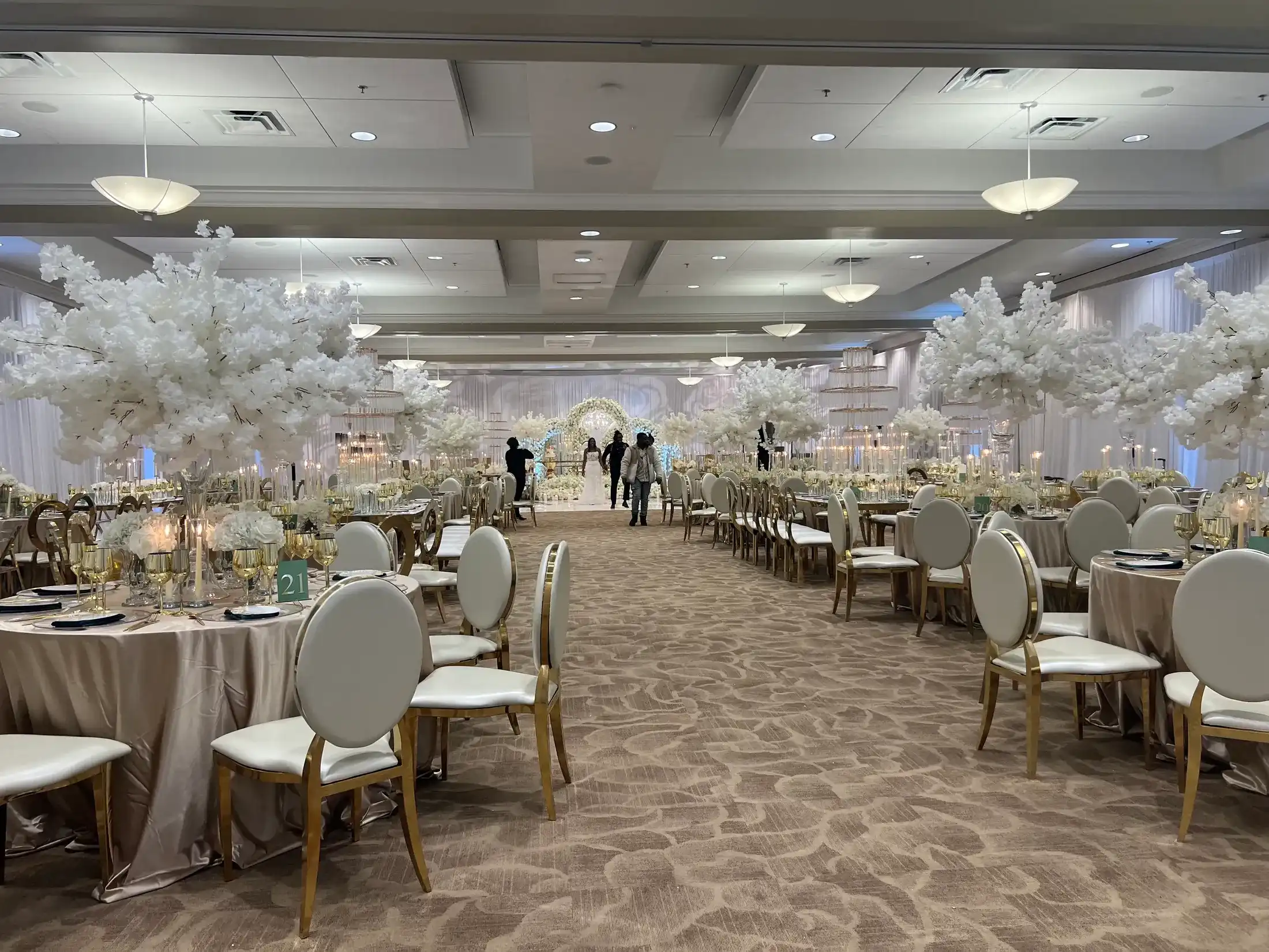 A grand ballroom wedding set with white blossom centerpieces, gold chairs and a floral arch at the end of the aisle