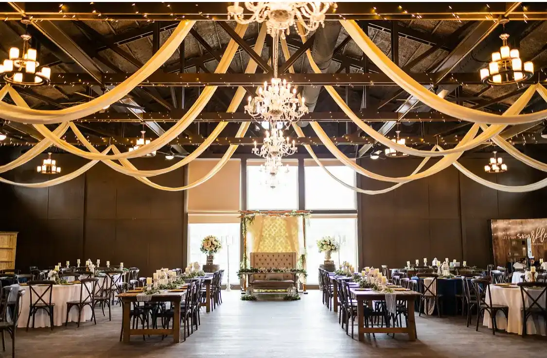 A reception hall with draped fabric overhead, crystal chandeliers and long wooden tables set for guests