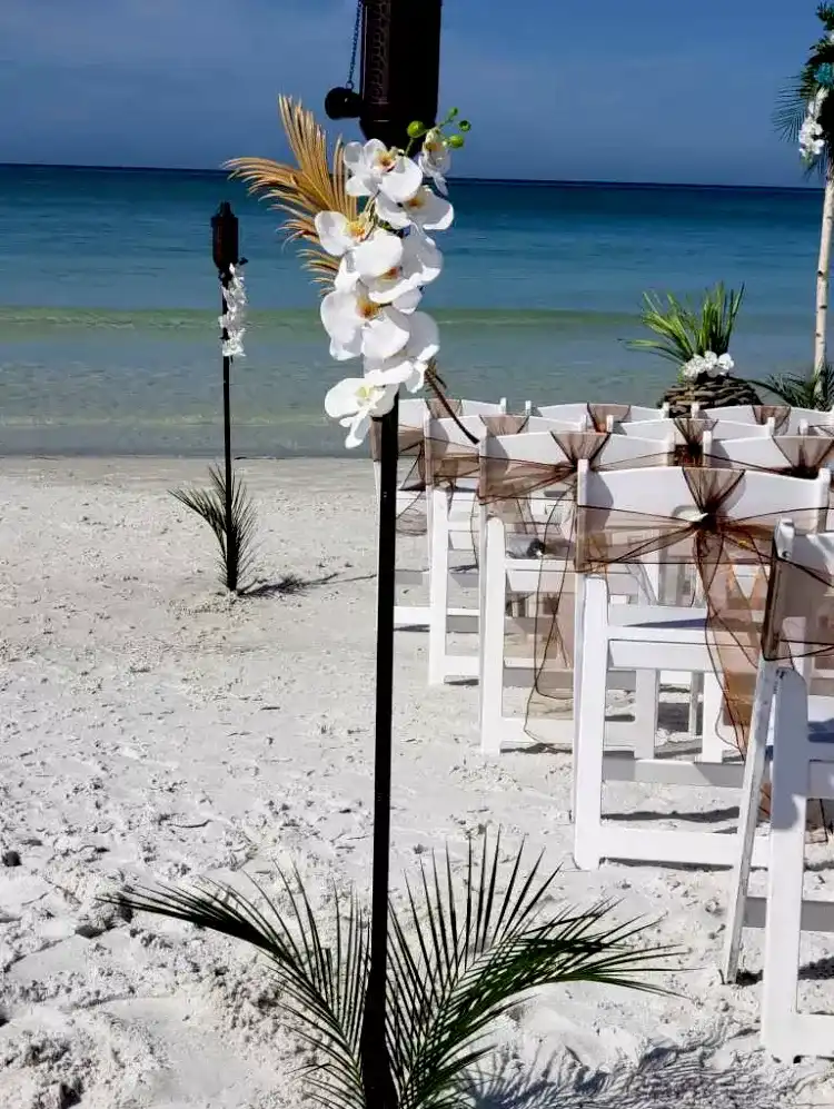 A beach ceremony aisle with white chairs, orchids and a tiki torch above the shoreline