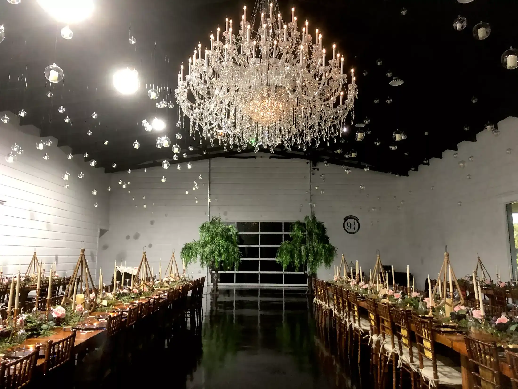 A long wedding reception hall at night with a large chandelier, hanging candle globes and tables set with pink florals