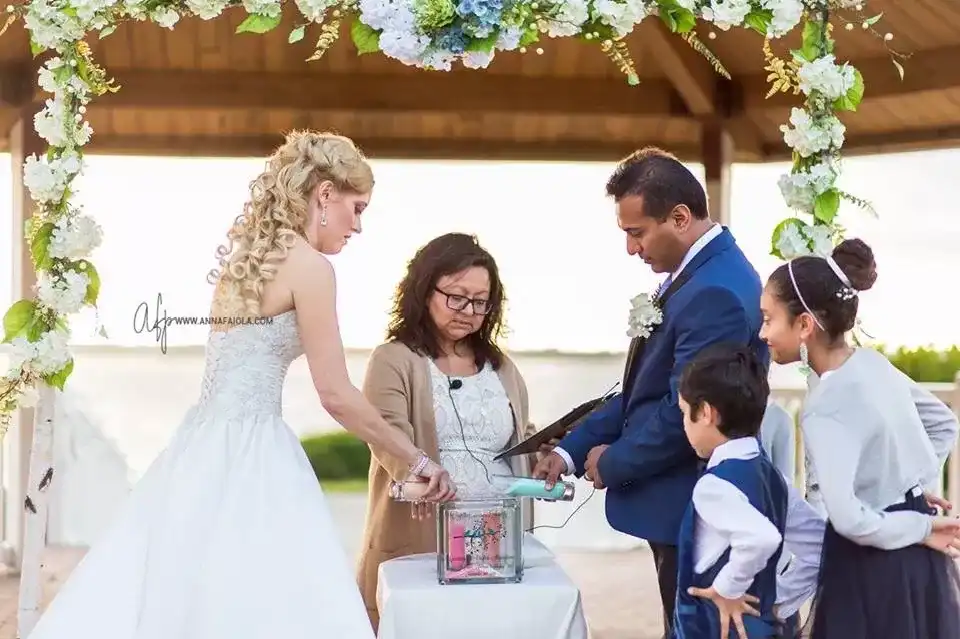 Rocío officiating a sand ceremony under a white hydrangea arch beside the water