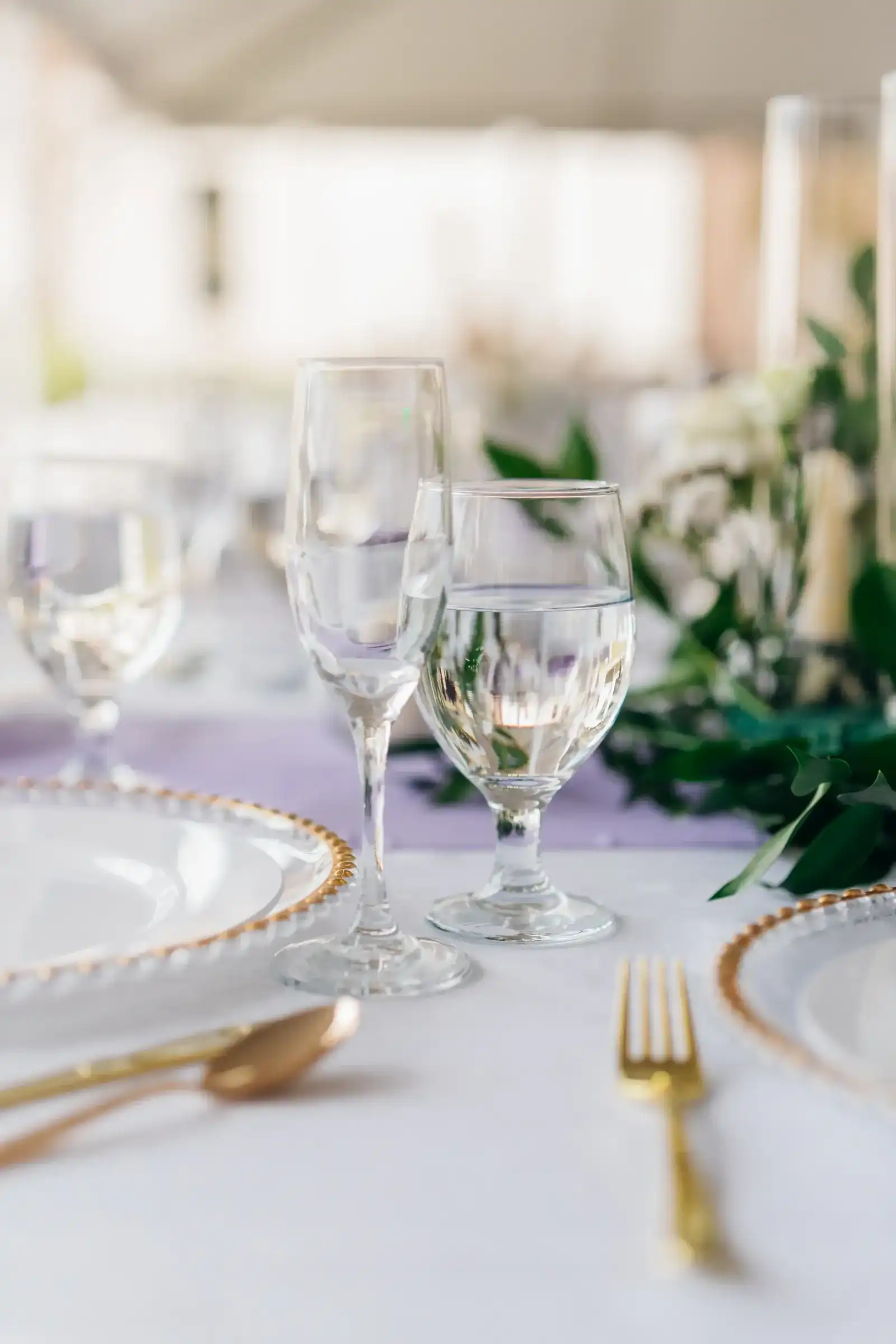 A place setting with a flute and goblet, a gold-rim charger, a gold fork and a lavender runner
