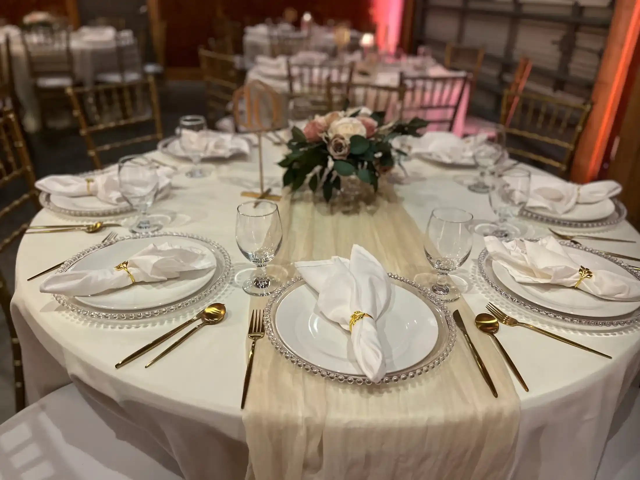 A round table with an ivory runner, beaded chargers, gold flatware and a blush rose centerpiece