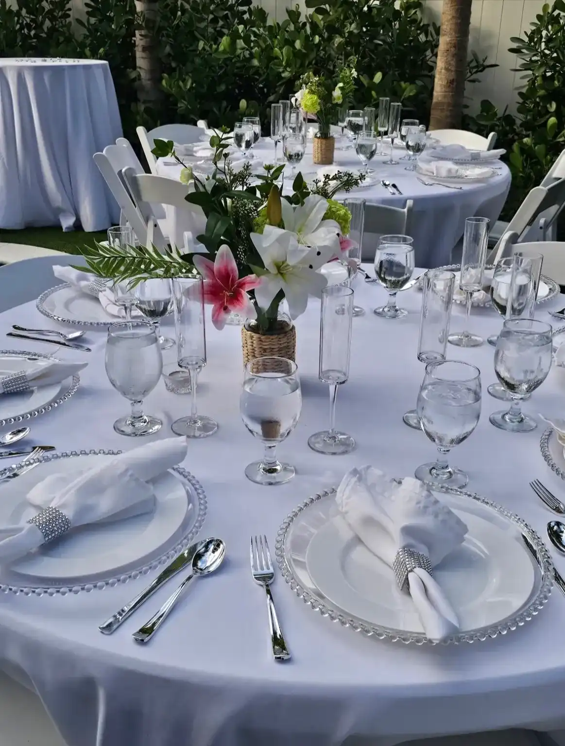 A round table with white linen, beaded glass chargers, folded napkins and a pink lily centerpiece