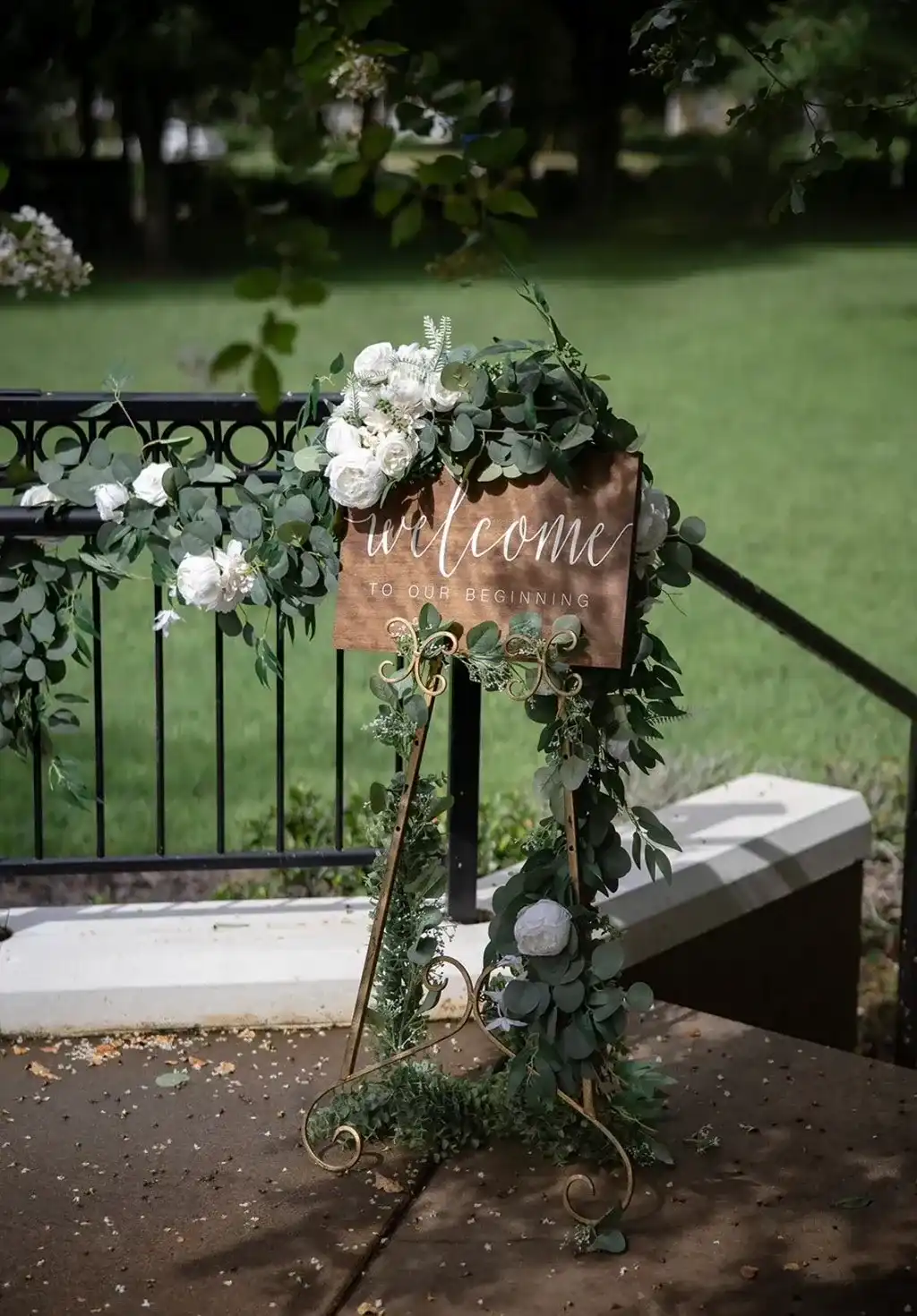 A wooden welcome sign on a gold easel wrapped in eucalyptus and white roses